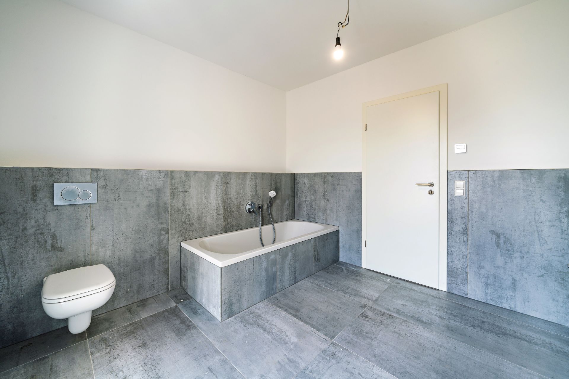 Modernes Bad mit Badewanne, Wandtoilette, grauen Fliesen und weißer Tür.