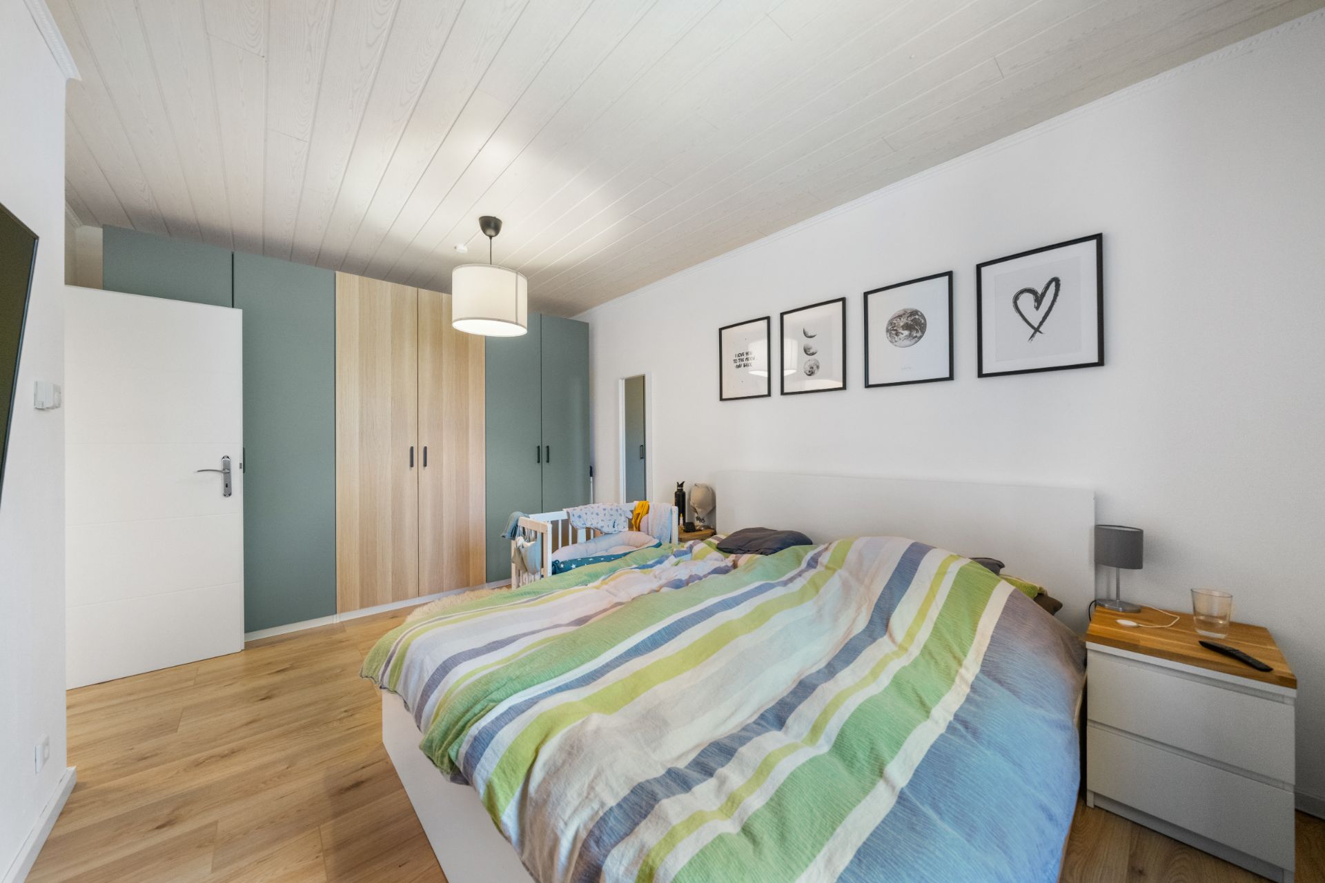 Schlafzimmer mit Doppelbett, bunter Bettdecke, Schrankwand und Bildern an der weißen Wand.