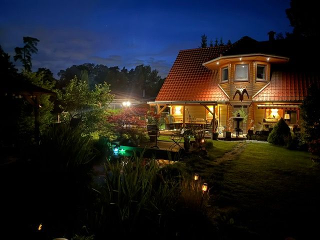 Garten mit beleuchtetem Haus und Teich bei Nacht, Bäume und Pflanzen im Vordergrund.