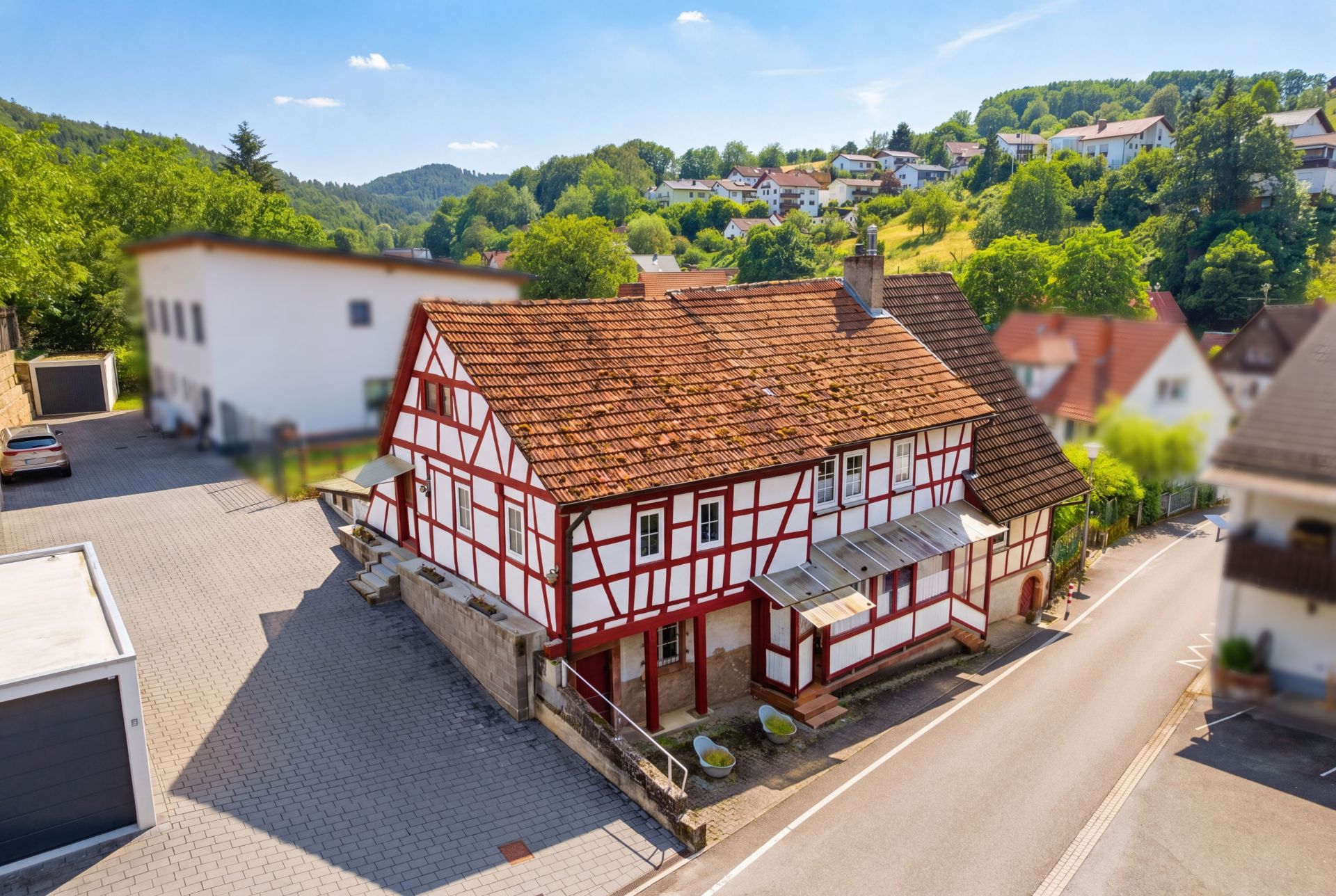 Fachwerkhaus mit rotem Dach an einer Straße, umgeben von grünen Hügeln und weiteren Häusern.