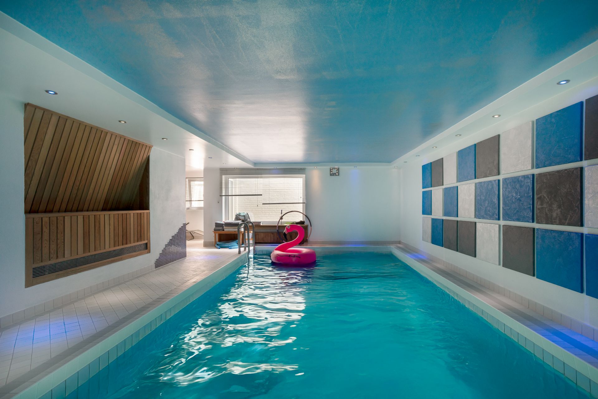 Heller Indoor-Pool mit Holzsauna, bunter Wand und aufblasbarem pinken Flamingo.