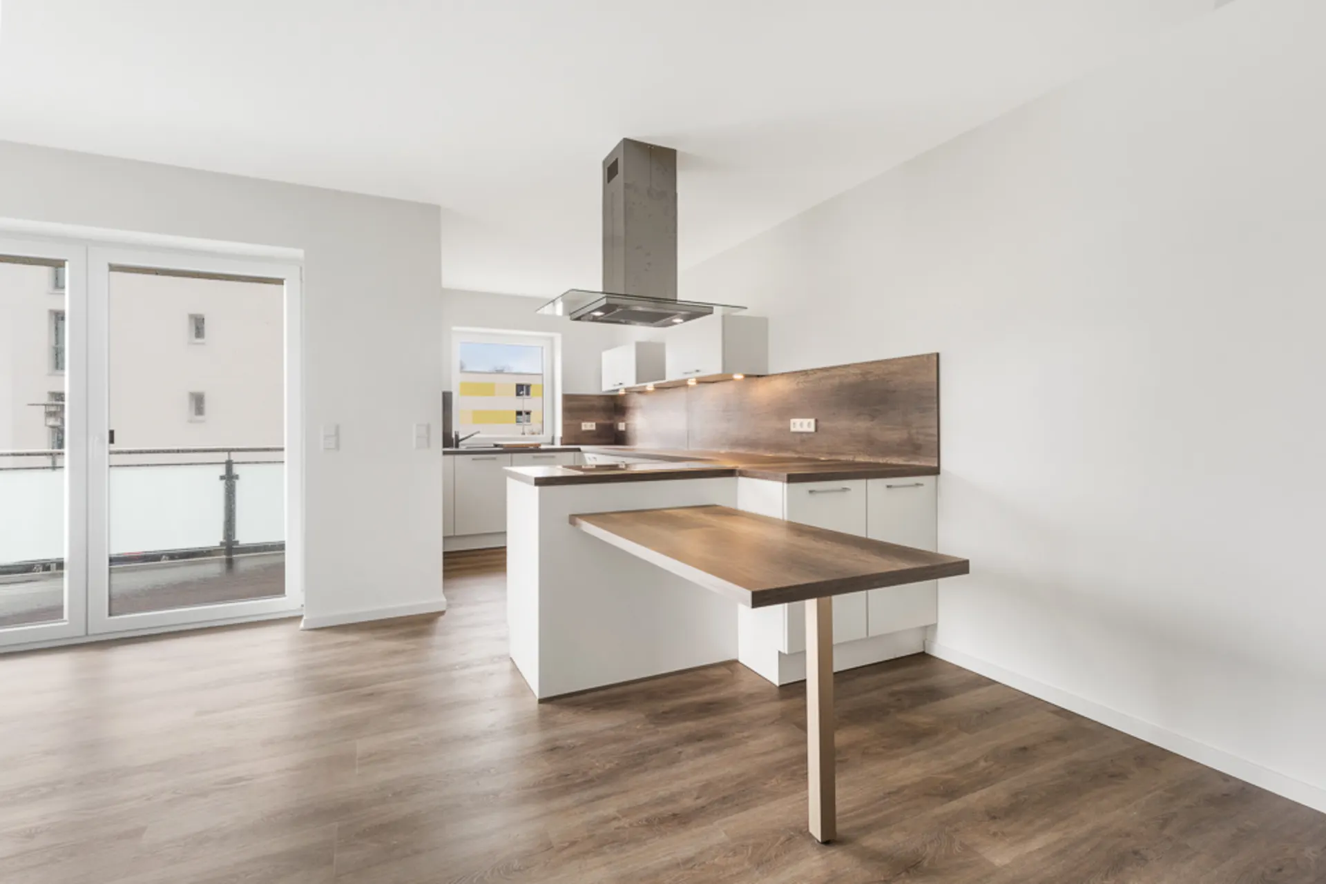 Moderne, helle Einbauküche mit Holzarbeitsplatte und Dunstabzugshaube, daneben Glastüren zum Balkon.