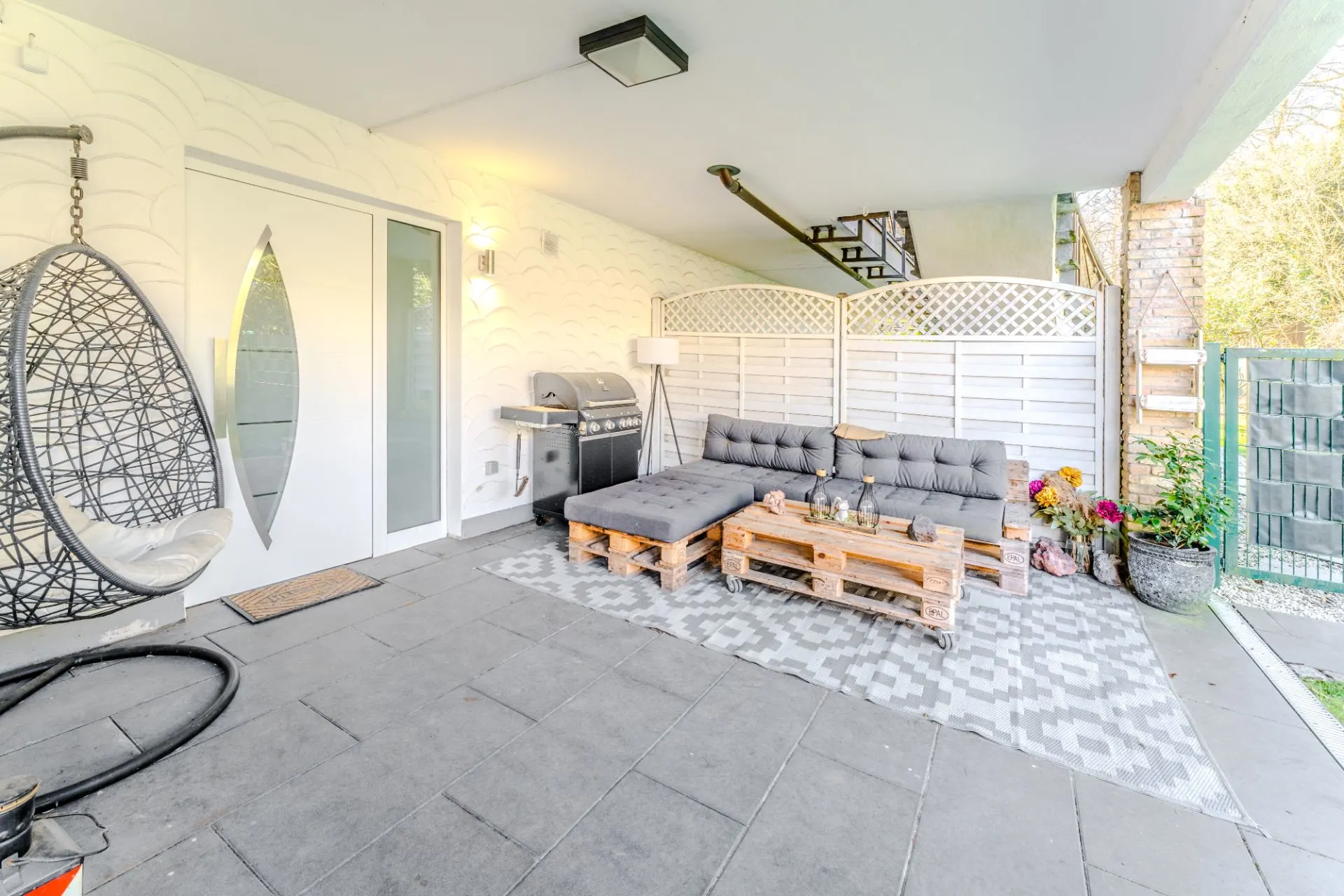Überdachte Terrasse mit Paletten-Sofa, Hängesessel, Teppich und Grill neben einer weißen Tür.