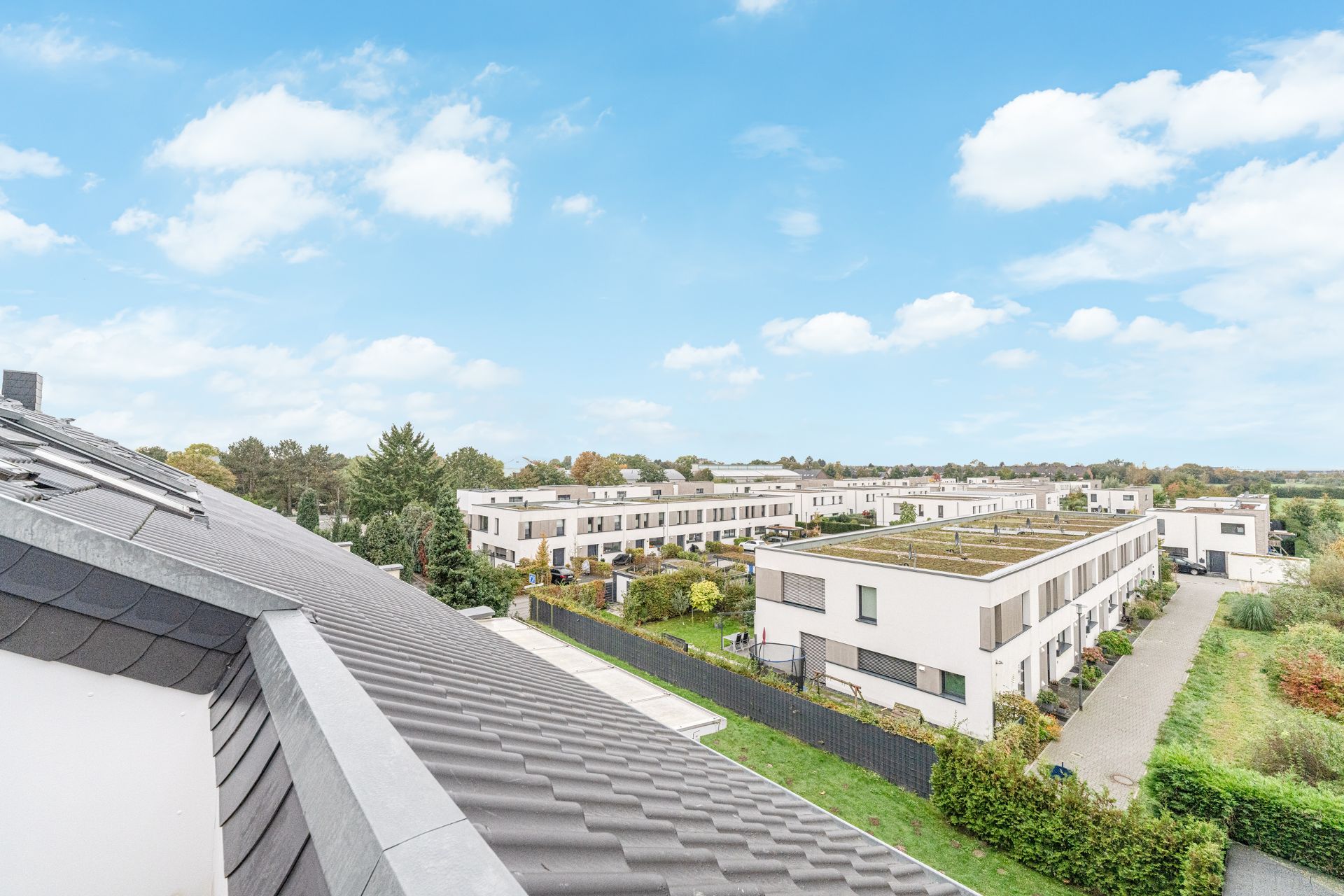 Blick auf moderne Reihenhäuser mit Flachdächern, Gärten und Grünanlagen bei blauem Himmel.
