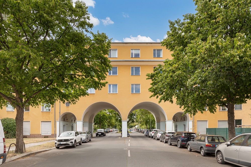 Gelbes Wohngebäude mit großen Torbögen über einer Straße, Autos parken am Straßenrand, Bäume säumen die Straße.
