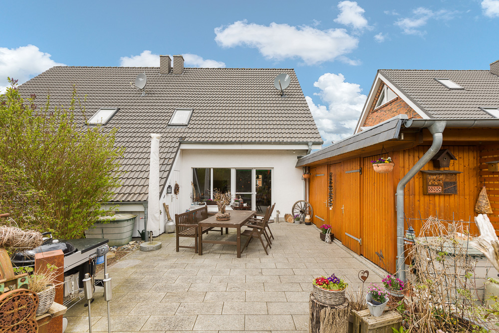 Gepflasterte Terrasse mit Holztisch und Stühlen, umgeben von Büschen und einem Nebengebäude.