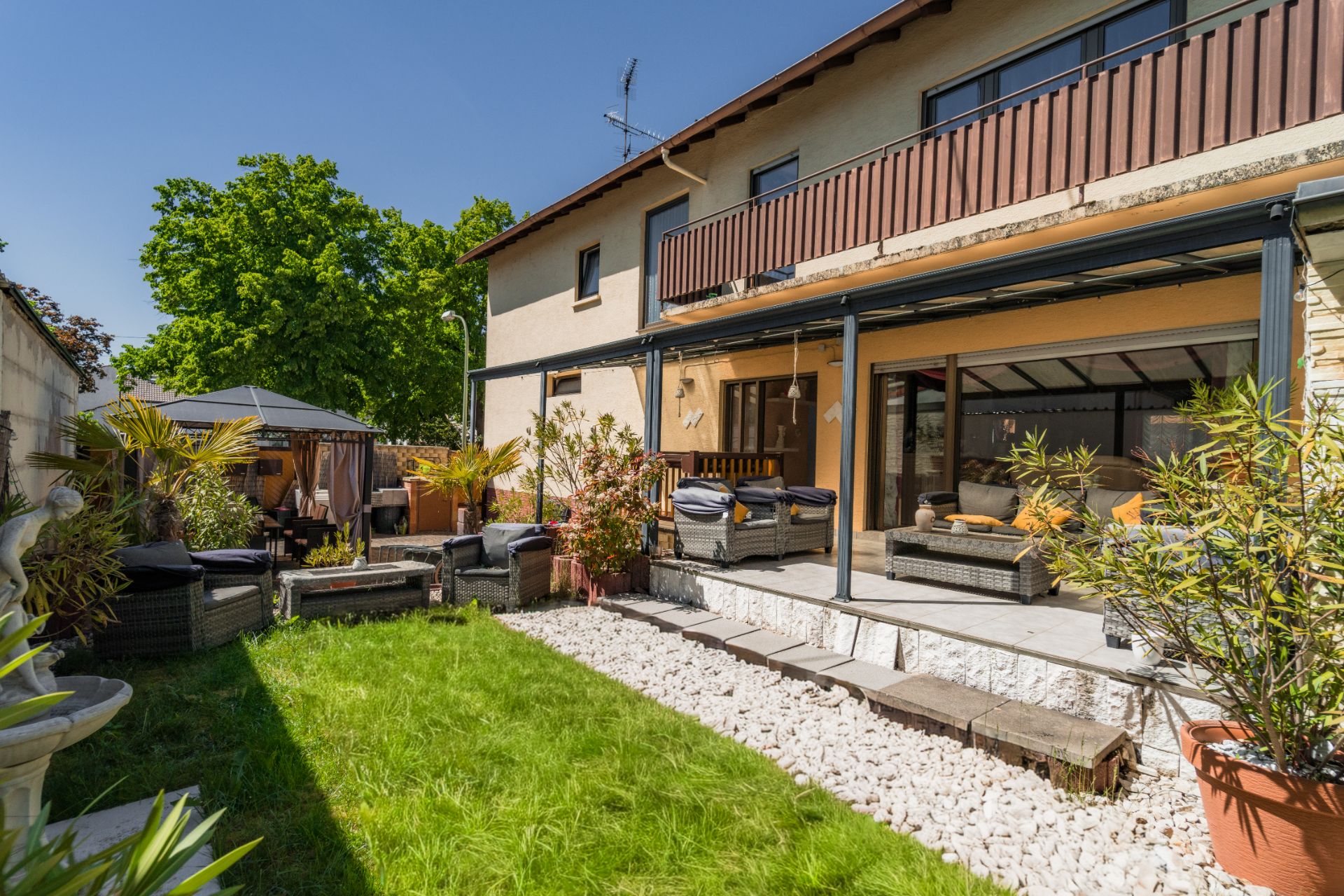 Garten mit Terrasse, Sitzmöbeln und Pflanzen vor einem zweistöckigen Wohnhaus an einem sonnigen Tag.