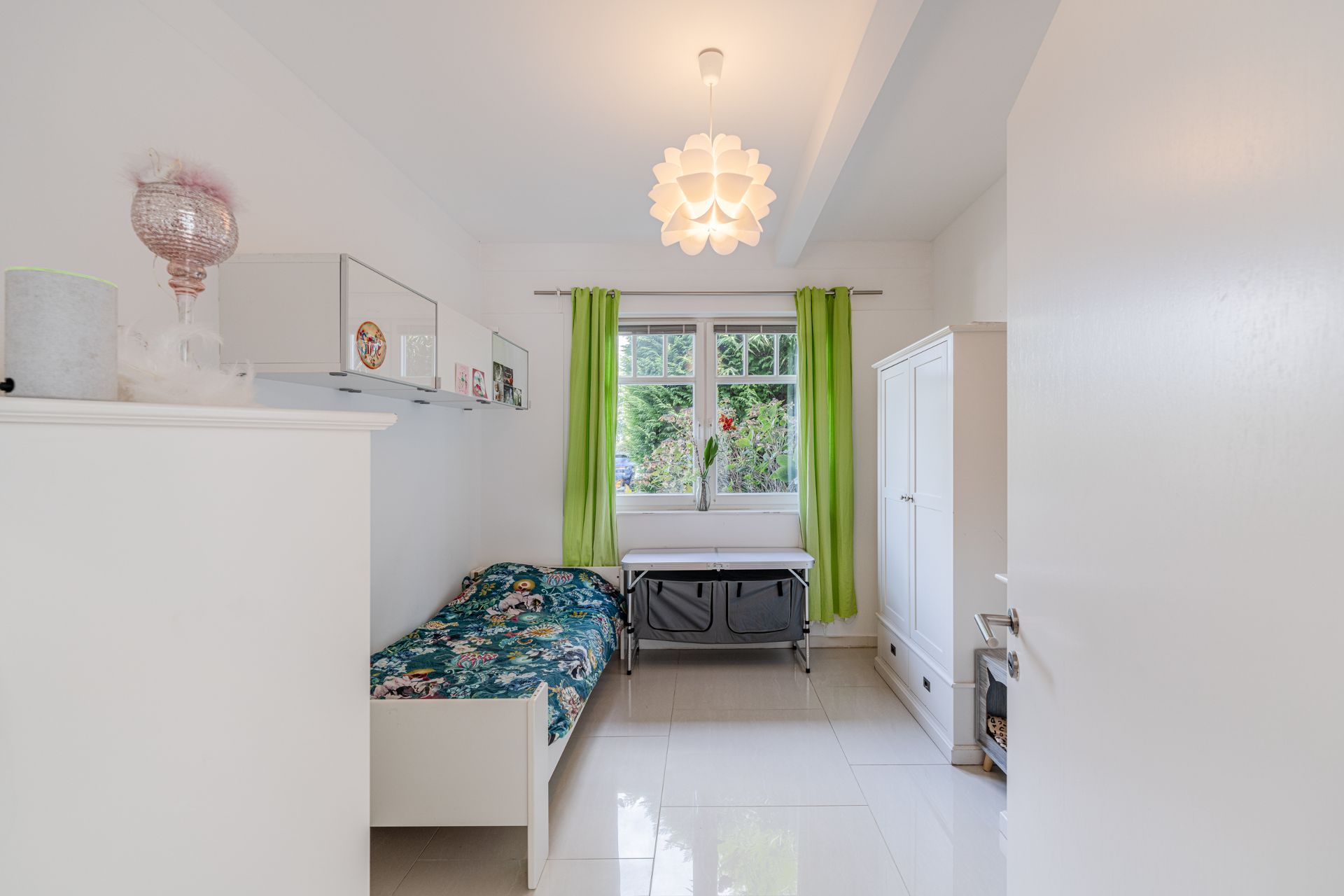 Helles Schlafzimmer mit Einzelbett, grünem Vorhang, Schrank und Fenster mit Blick ins Grüne.
