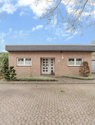 Einstöckiges Haus mit Flachdach, Backsteinfassade und großen Fenstern, vor einem gepflasterten Hof.