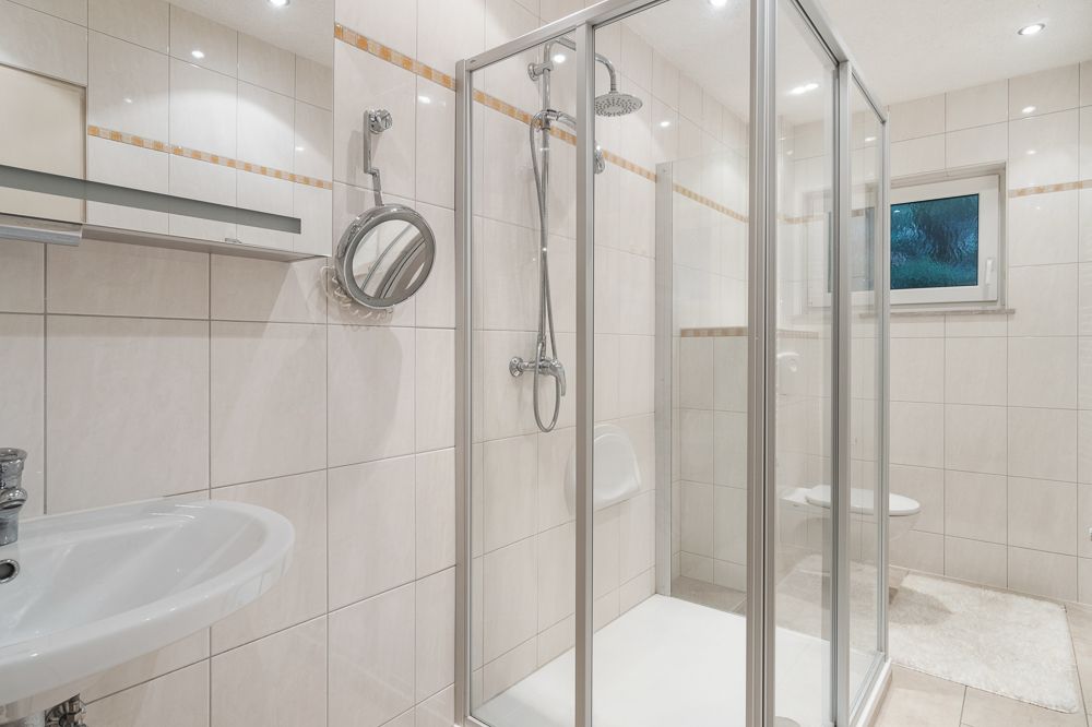 Helles Bad mit Dusche, Waschbecken, Spiegel, Fenster und Toilette auf gefliesten Wänden.