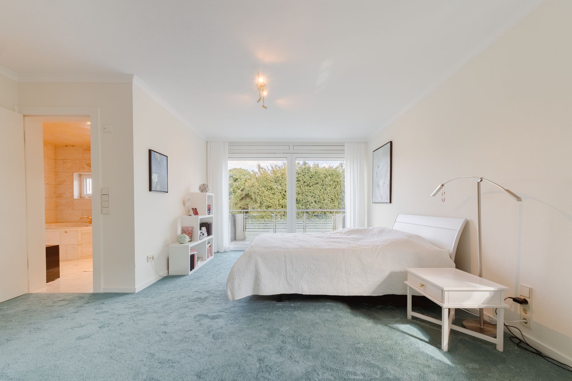 Helles Schlafzimmer mit Doppelbett, Nachttisch und Zugang zum Balkon, Teppichboden und angrenzendem Badezimmer.