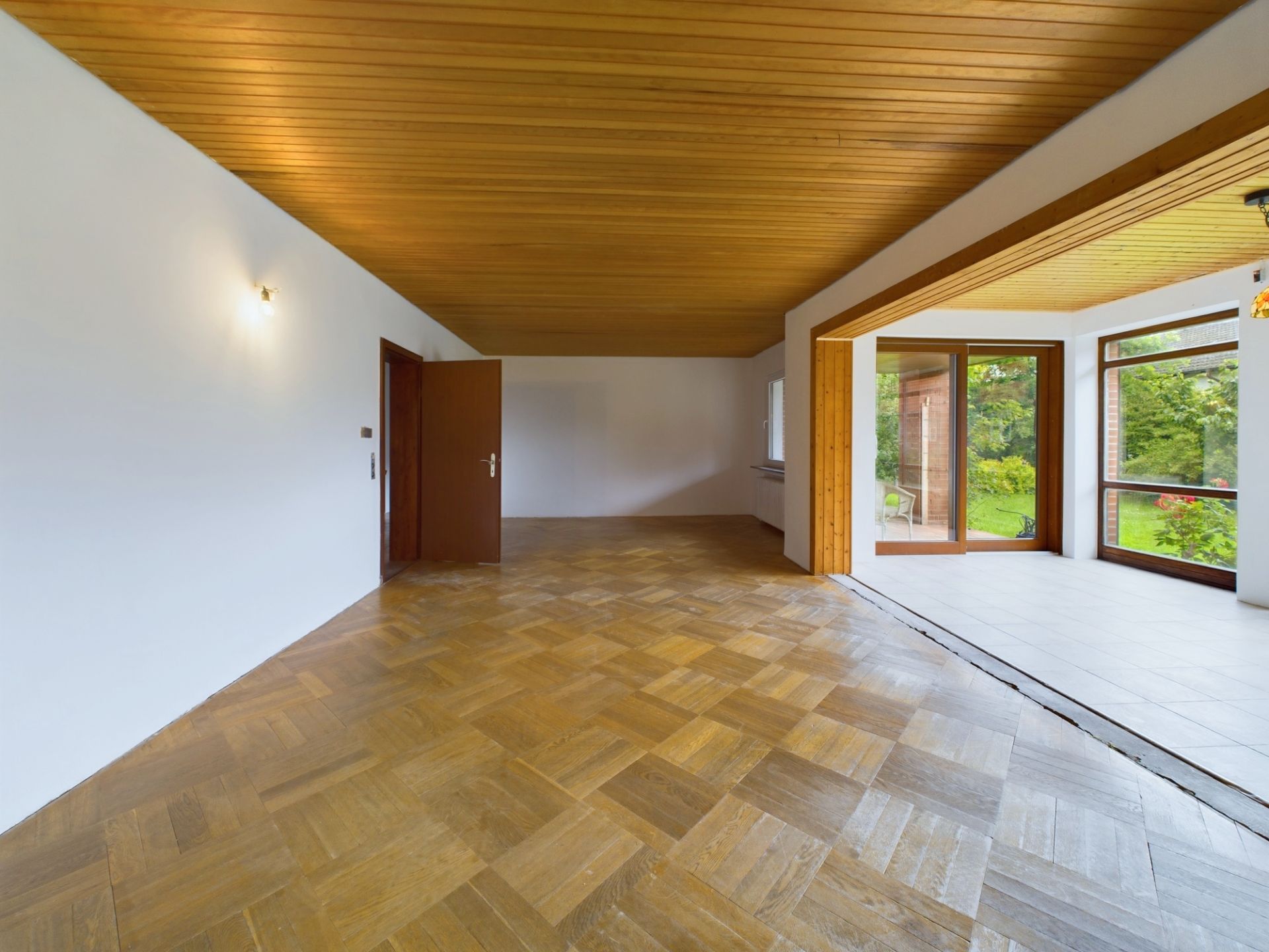 Leeres Zimmer mit Parkettboden, Holzdecke und großen Fenstern zum Garten.
