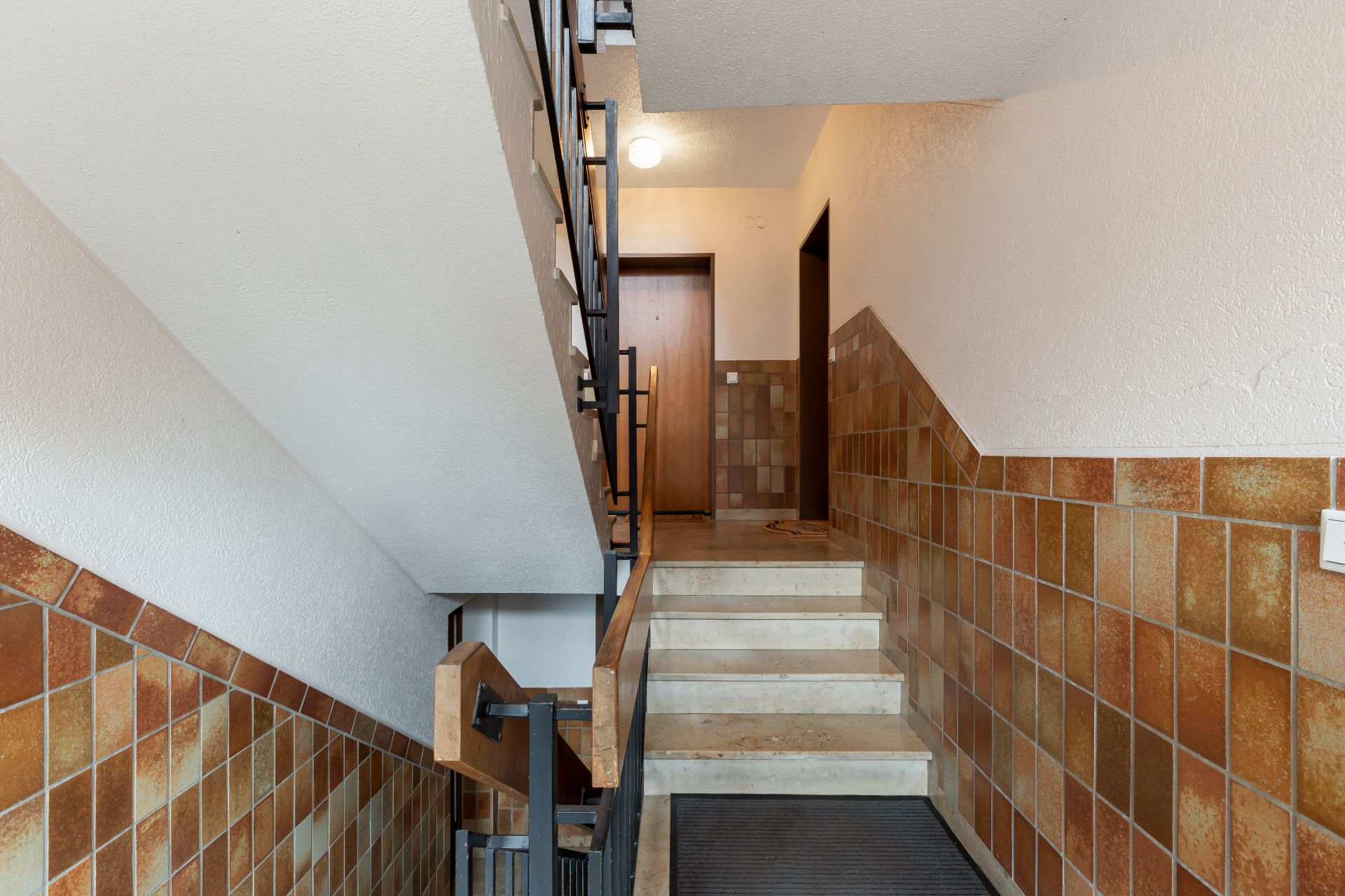 Treppenhaus mit braunen Fliesen, Handlauf und Tür am oberen Ende der Treppe.