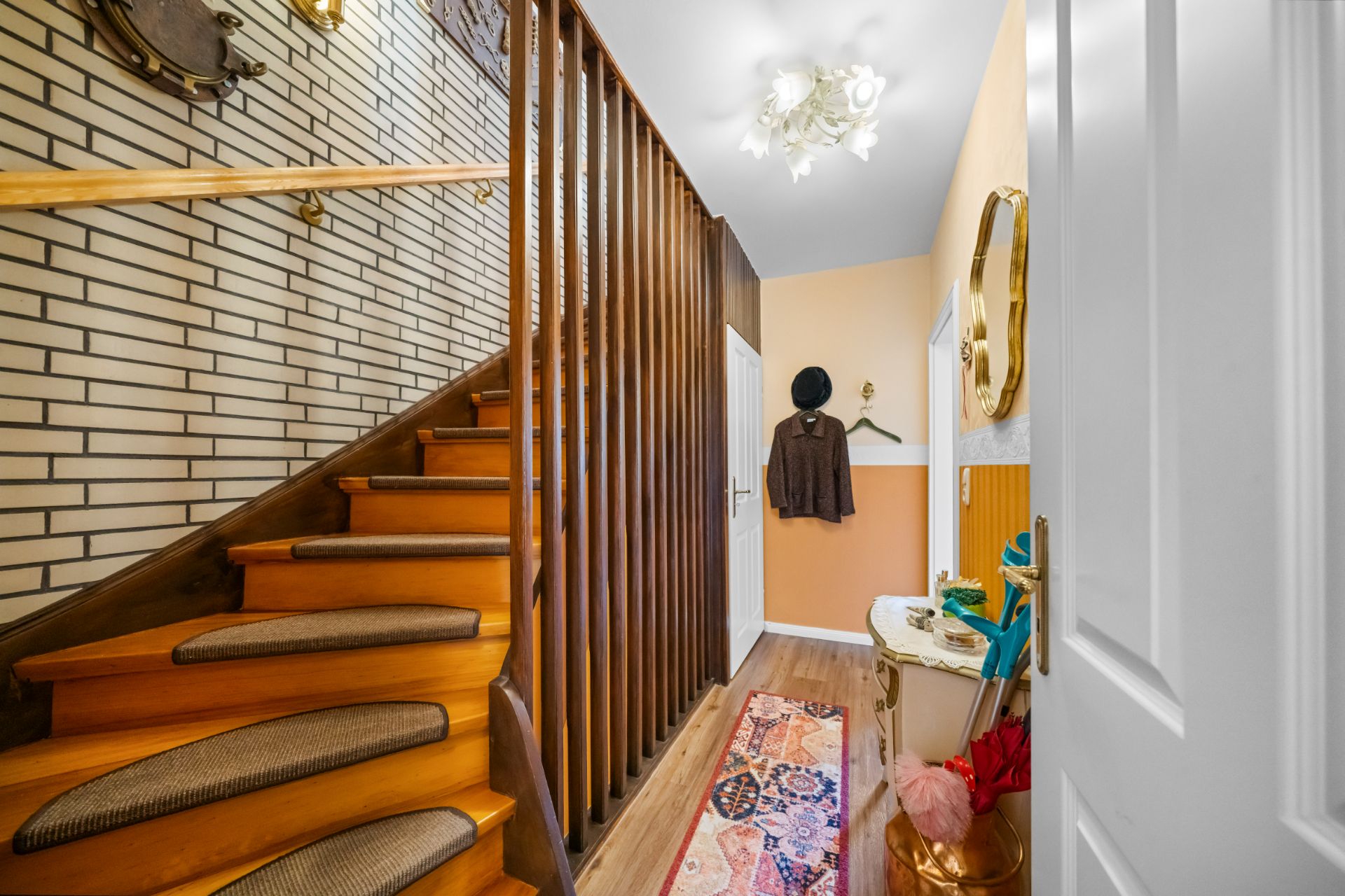 Flur mit Holztreppe, Garderobe, Spiegel und bunter Teppichläufer.