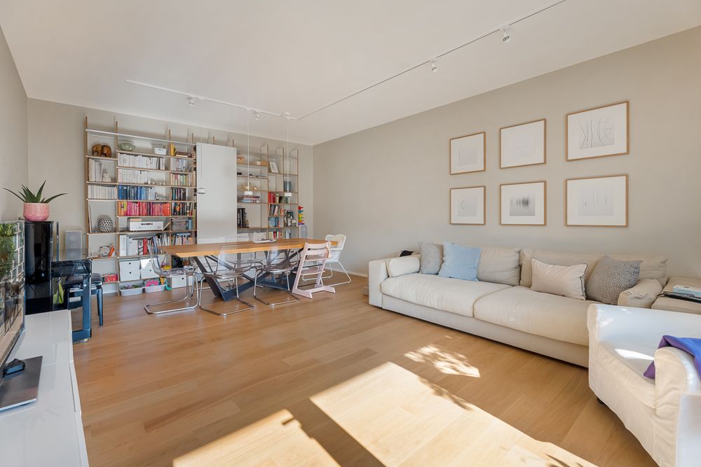 Helles Wohnzimmer mit Bücherregal, langem Tisch, weißem Sofa und an der Wand hängenden Bildern.