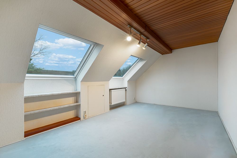 Leerer Dachzimmer mit zwei großen Dachfenstern, Holzdecke und Regal an der linken Wand.