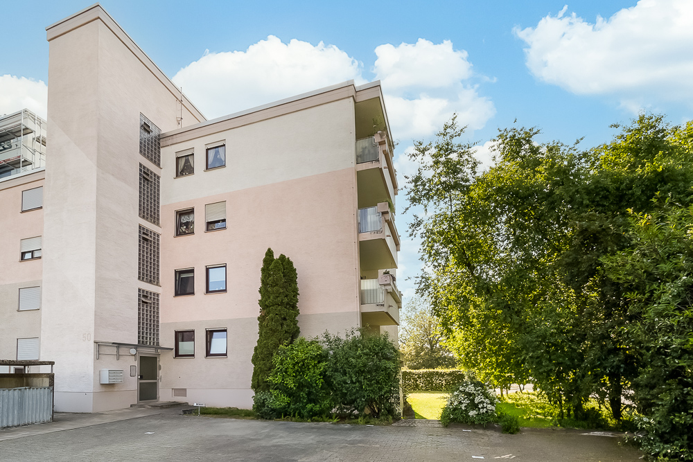 Mehrfamilienhaus mit Balkonen, davor Bäume und Sträucher, blauer Himmel im Hintergrund.