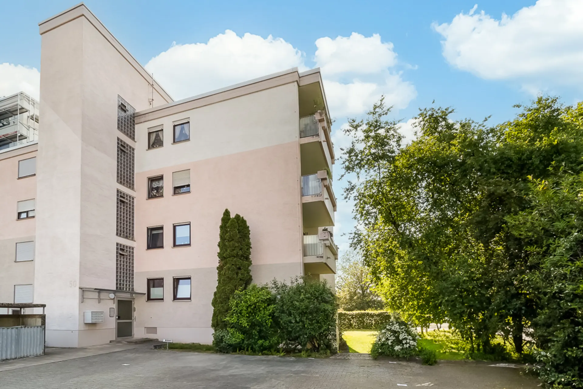 Mehrfamilienhaus mit Balkonen, davor Bäume und Sträucher, blauer Himmel im Hintergrund.