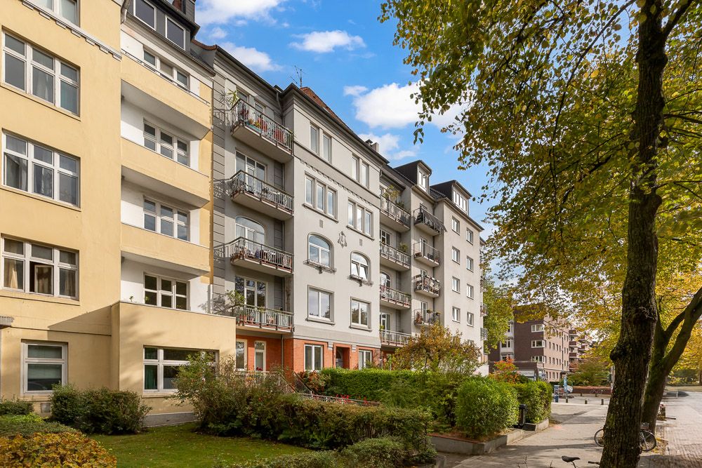 Mehrfamilienhaus mit Balkonen, Bäumen und Gehweg an einer ruhigen Straße.