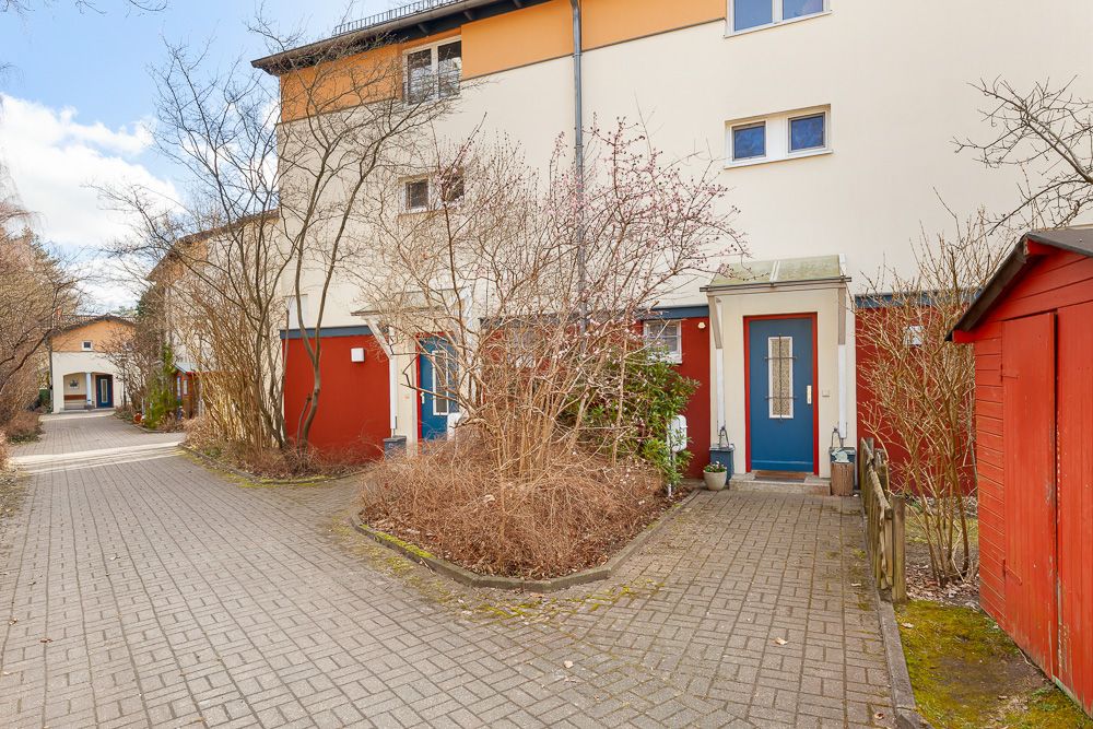 Eingang eines Mehrfamilienhauses mit blau-roten Türen und umliegenden Büschen im Vorgarten.