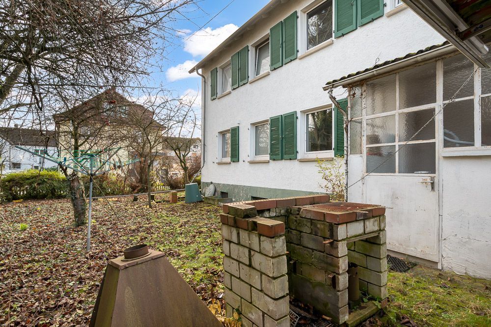 Garten mit gemauertem Grill und Wäscheleinen, neben einem weißen Wohnhaus mit grünen Fensterläden.