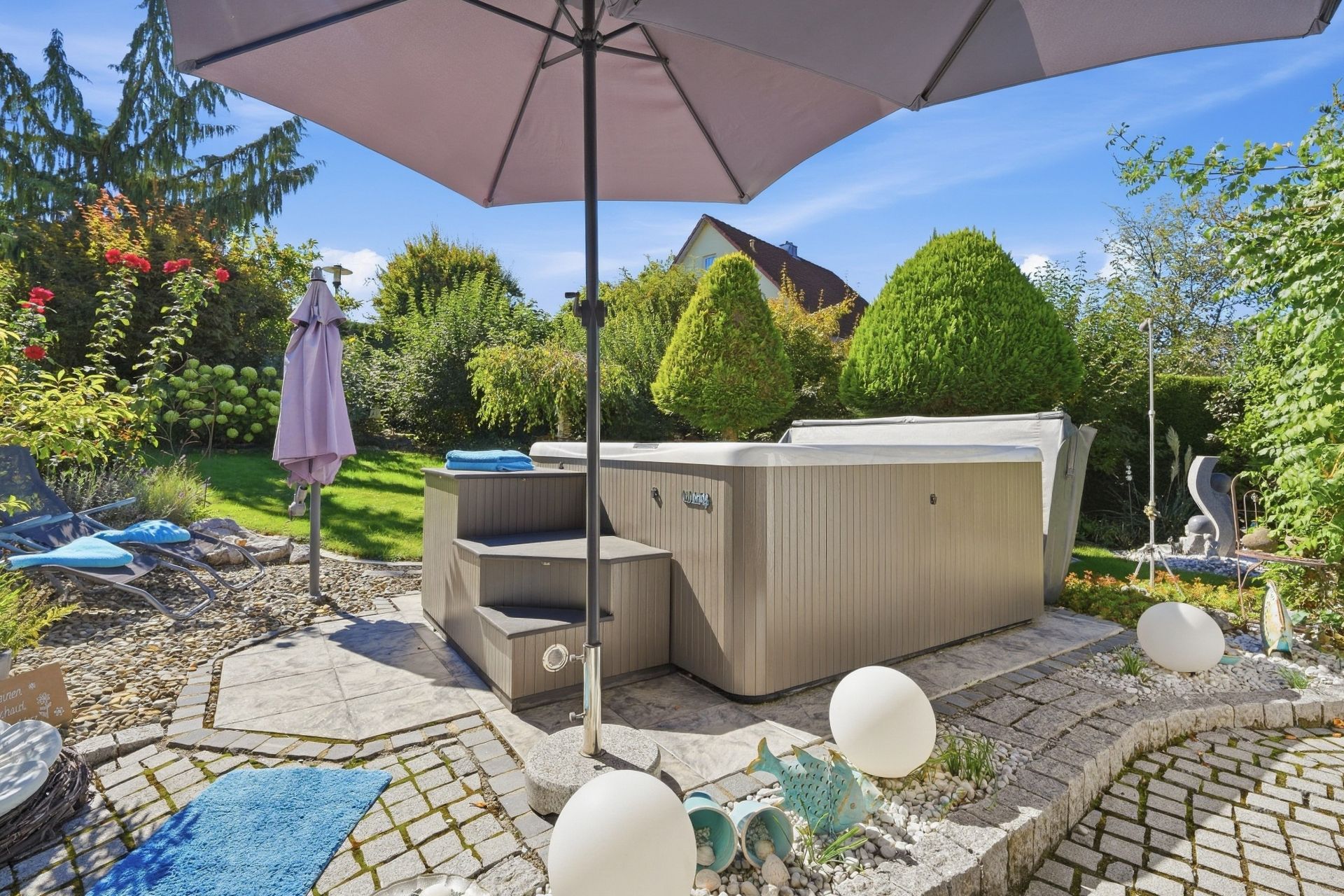 Außenbereich mit Whirlpool, Sonnenschirm, Liegen und Garten im Hintergrund.
