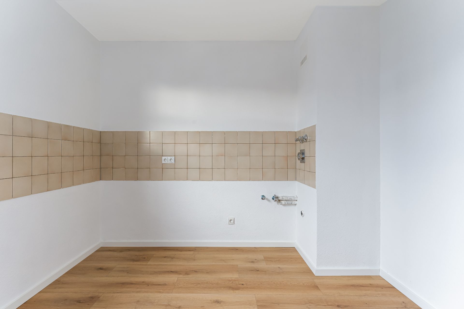 Leerer Raum mit beigem Fliesenspiegel und hellem Holzboden, Steckdosen an der Wand.