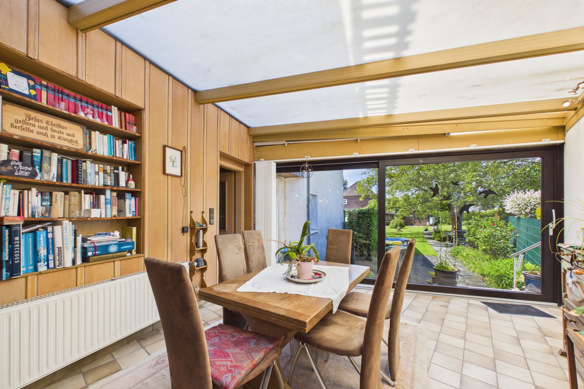 Essbereich mit Holzwand, Bücherregal und Blick durch große Fenster in einen Garten.