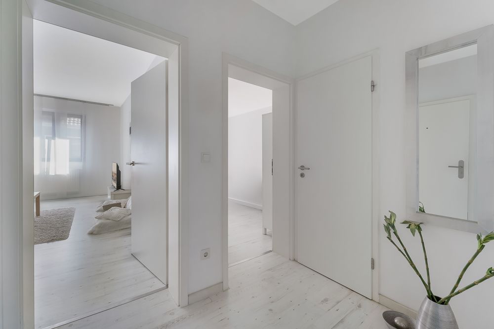 Heller Flur mit weißen Türen, Spiegel und Pflanze in einer Vase, Blick ins Wohnzimmer.
