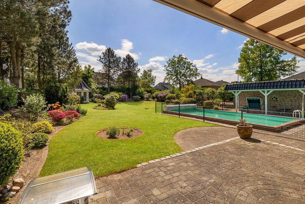 Garten mit Rasenfläche, Blumenbeeten, Pool mit Zaun und überdachter Terrasse bei sonnigem Wetter.