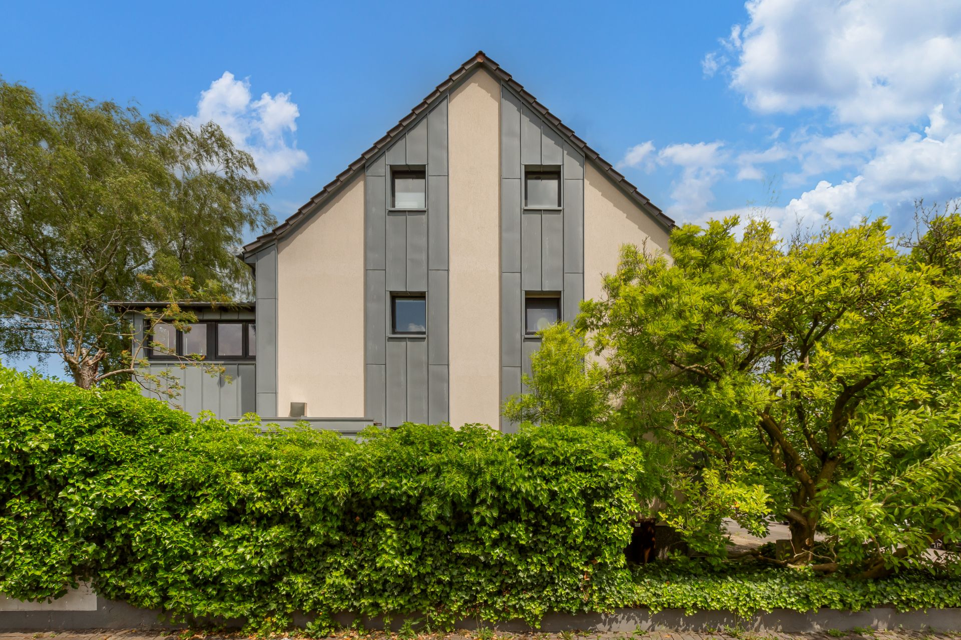 Vorderseite eines modernen Wohnhauses mit grauer Fassade und viel Grün im Vordergrund.