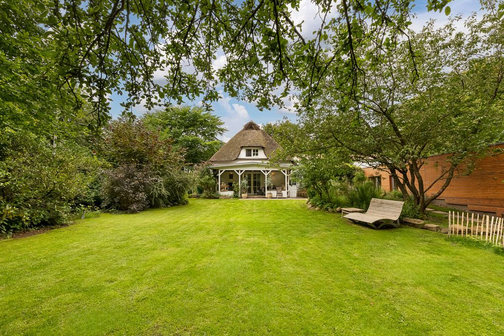 Ein Haus mit Reetdach steht in einem großen, grünen Garten mit Bäumen und Liege im Vordergrund.