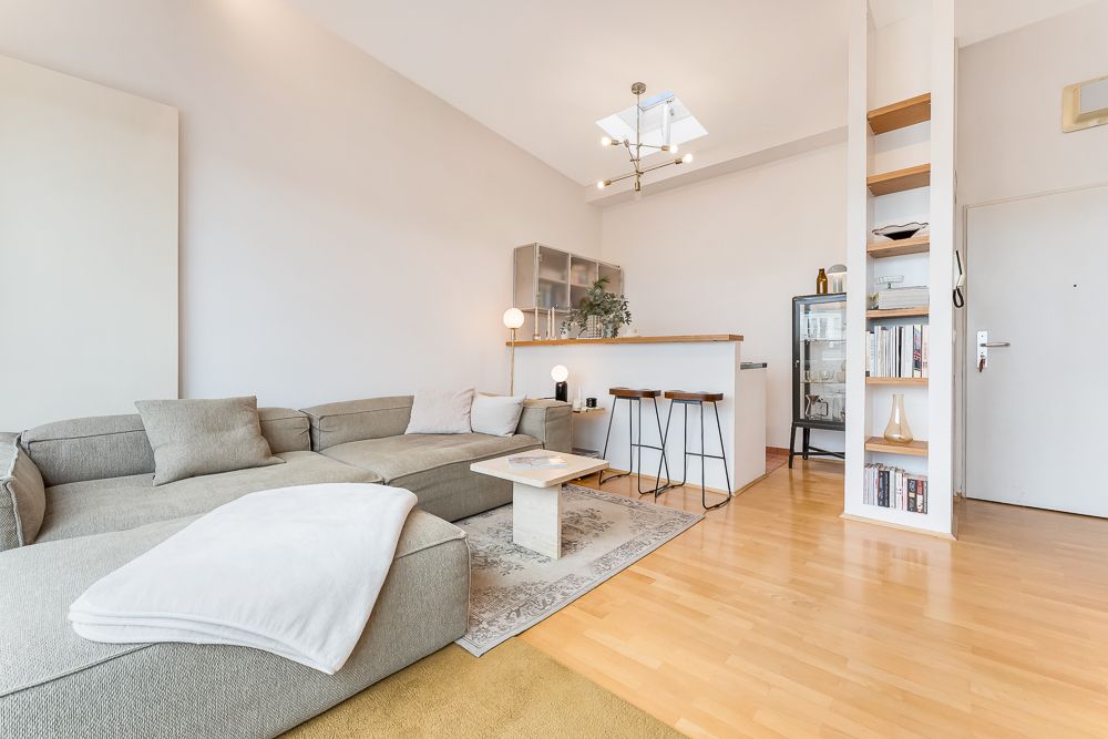 Modernes Wohnzimmer mit grauem Ecksofa, Couchtisch, offener Küche und Regal auf Holzboden.