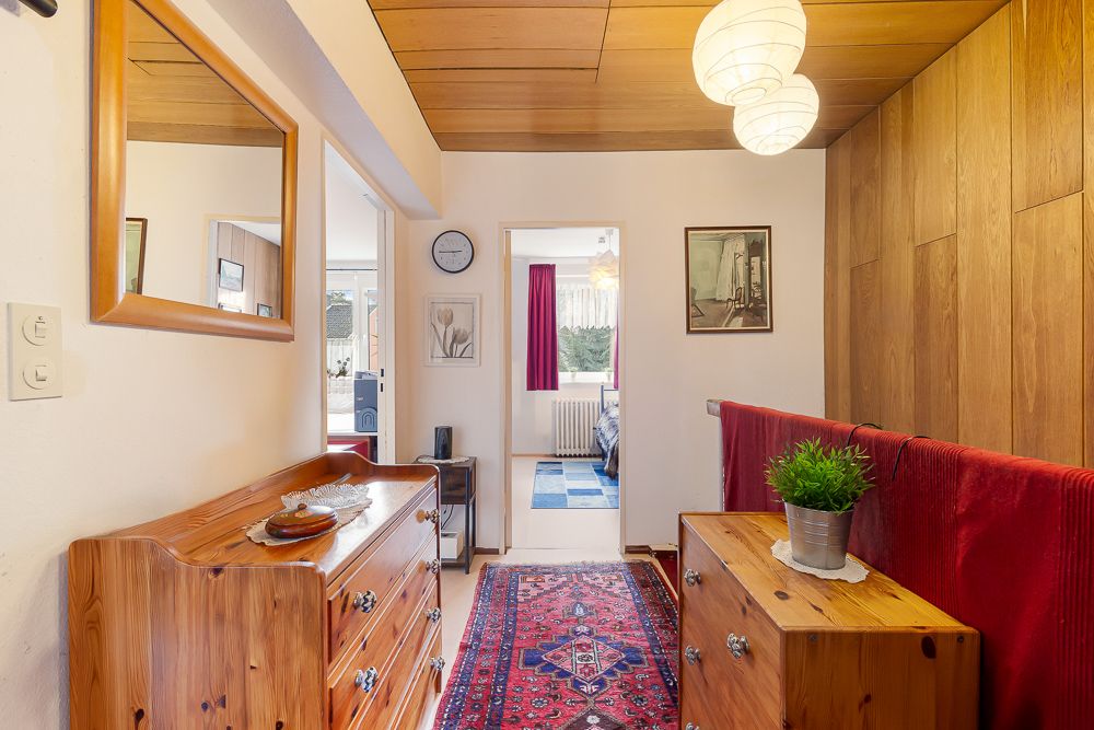 Heller Flur mit Holzmöbeln, Teppich, Spiegel und Blick in ein Zimmer mit roten Vorhängen.
