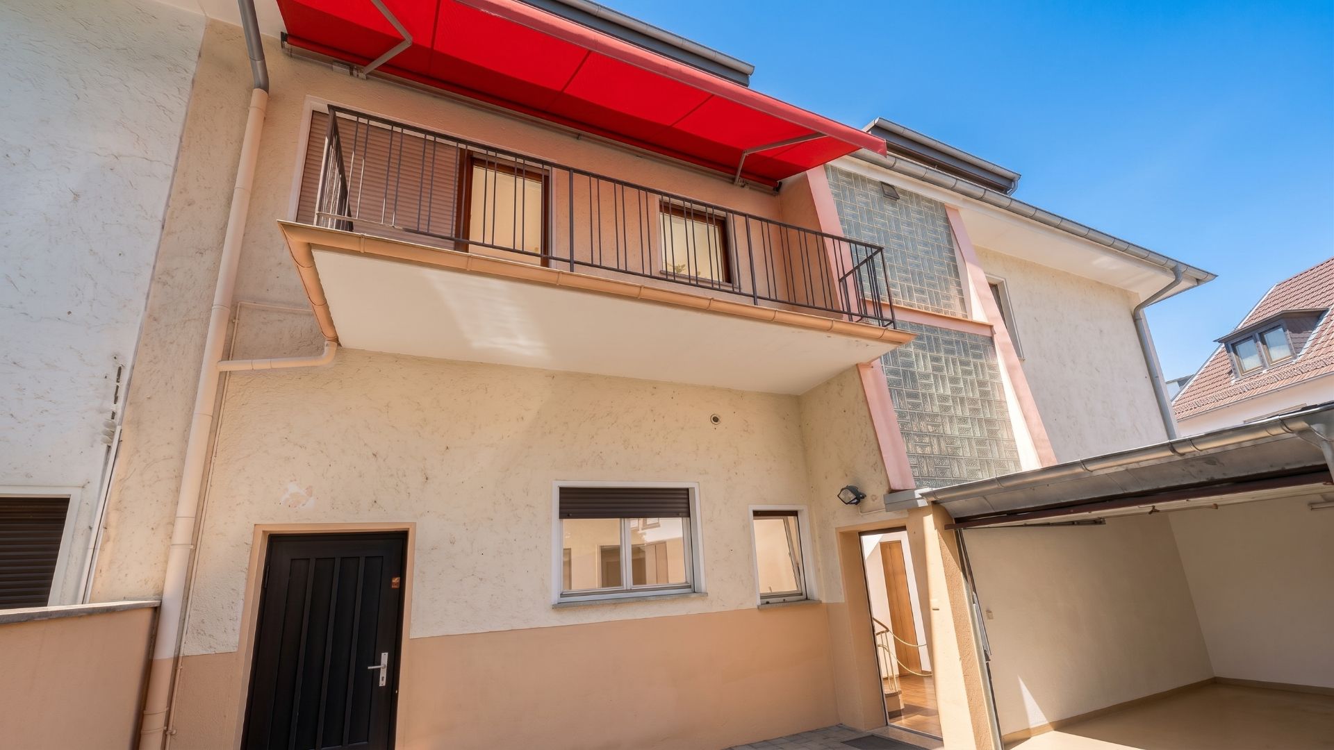 Zweistöckiges Wohnhaus mit Balkon, roter Markise und offenem Hofbereich.