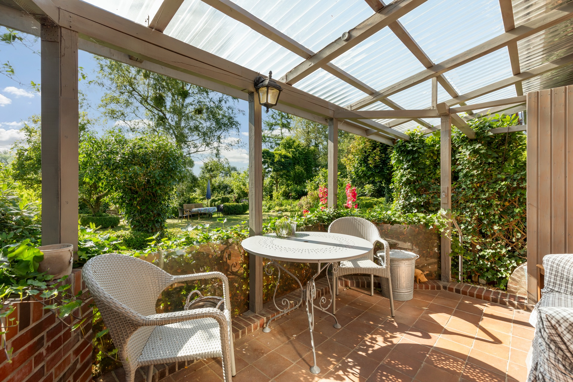 Überdachte Terrasse mit zwei weißen Stühlen und rundem Tisch, umgeben von viel Grün.