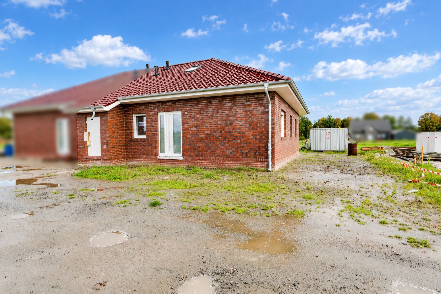 Backsteinwohnhaus mit rotem Dach, steht auf teilweise unbefestigtem Grundstück bei klarem Himmel.