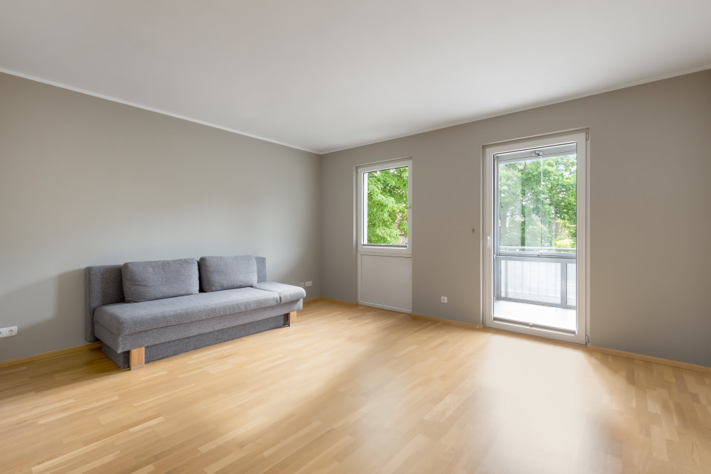 Helles Wohnzimmer mit Holzboden, grauem Sofa und großen Fenstern zum Balkon.