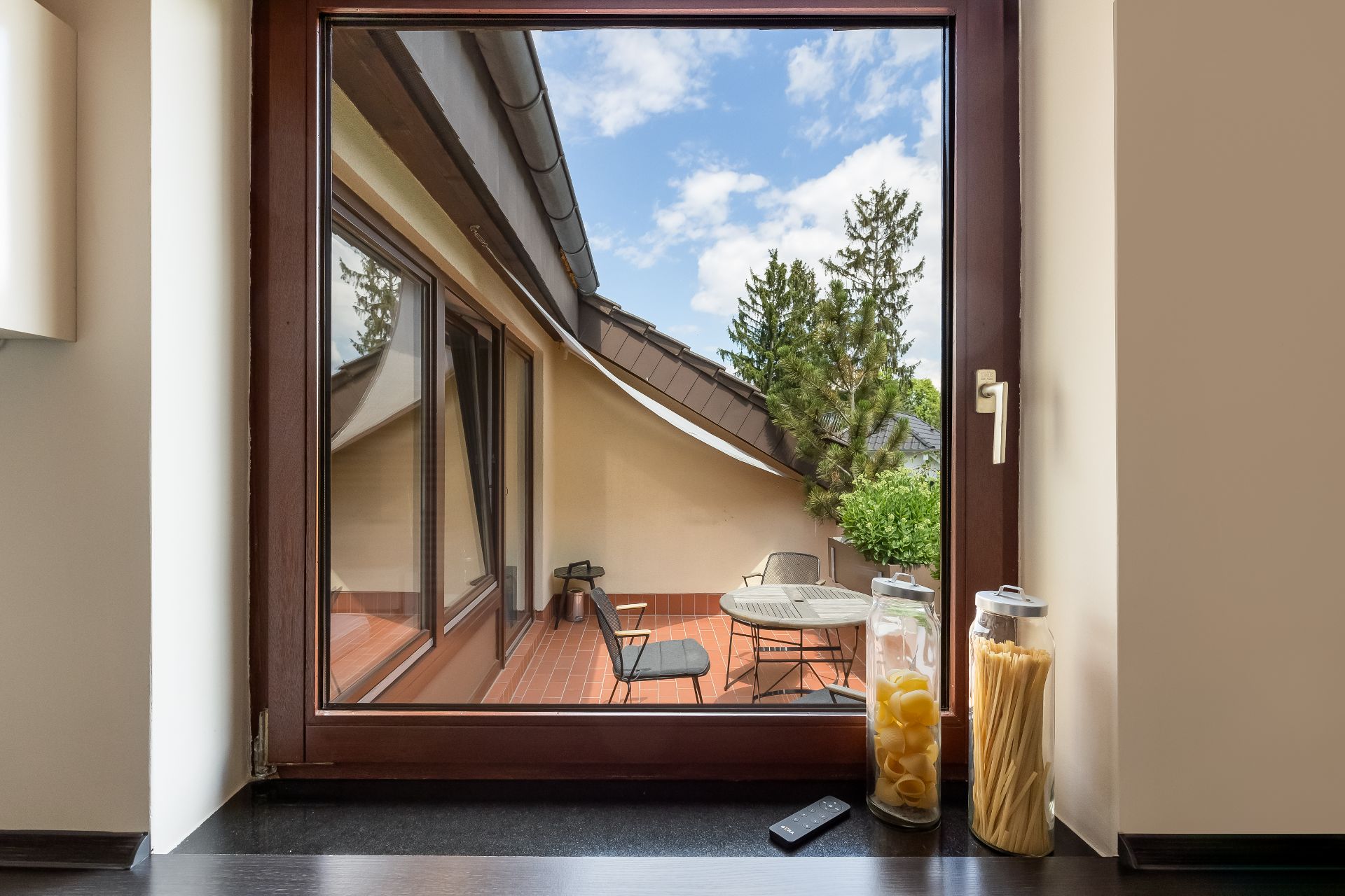 Blick durch Fenster auf Terrasse mit Tisch, Stühlen und Pflanzen, zwei Vorratsgläser auf der Fensterbank.