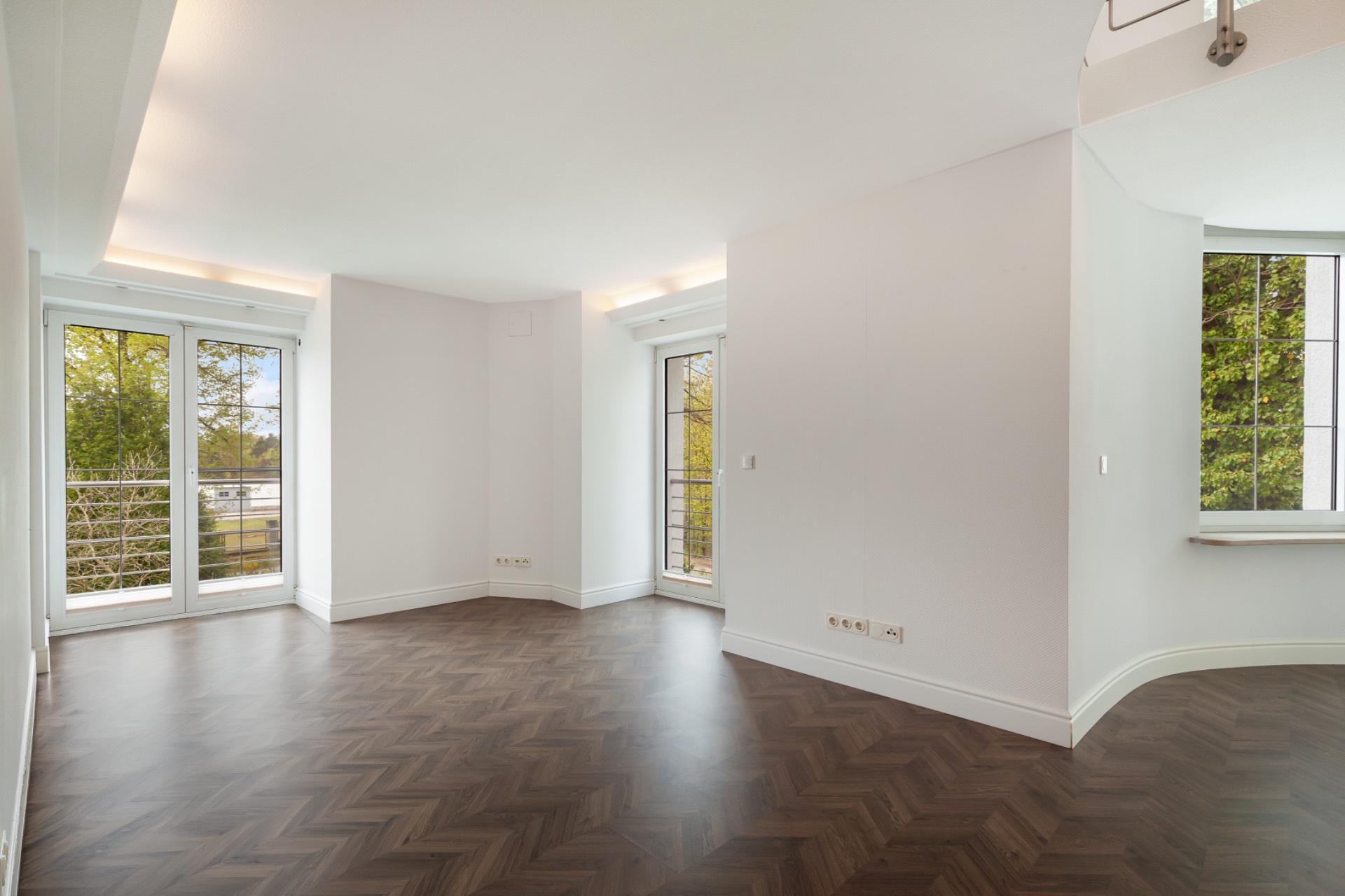 Heller Raum mit dunklem Holzboden, weißen Wänden und großen Fenstern mit Blick ins Grüne.