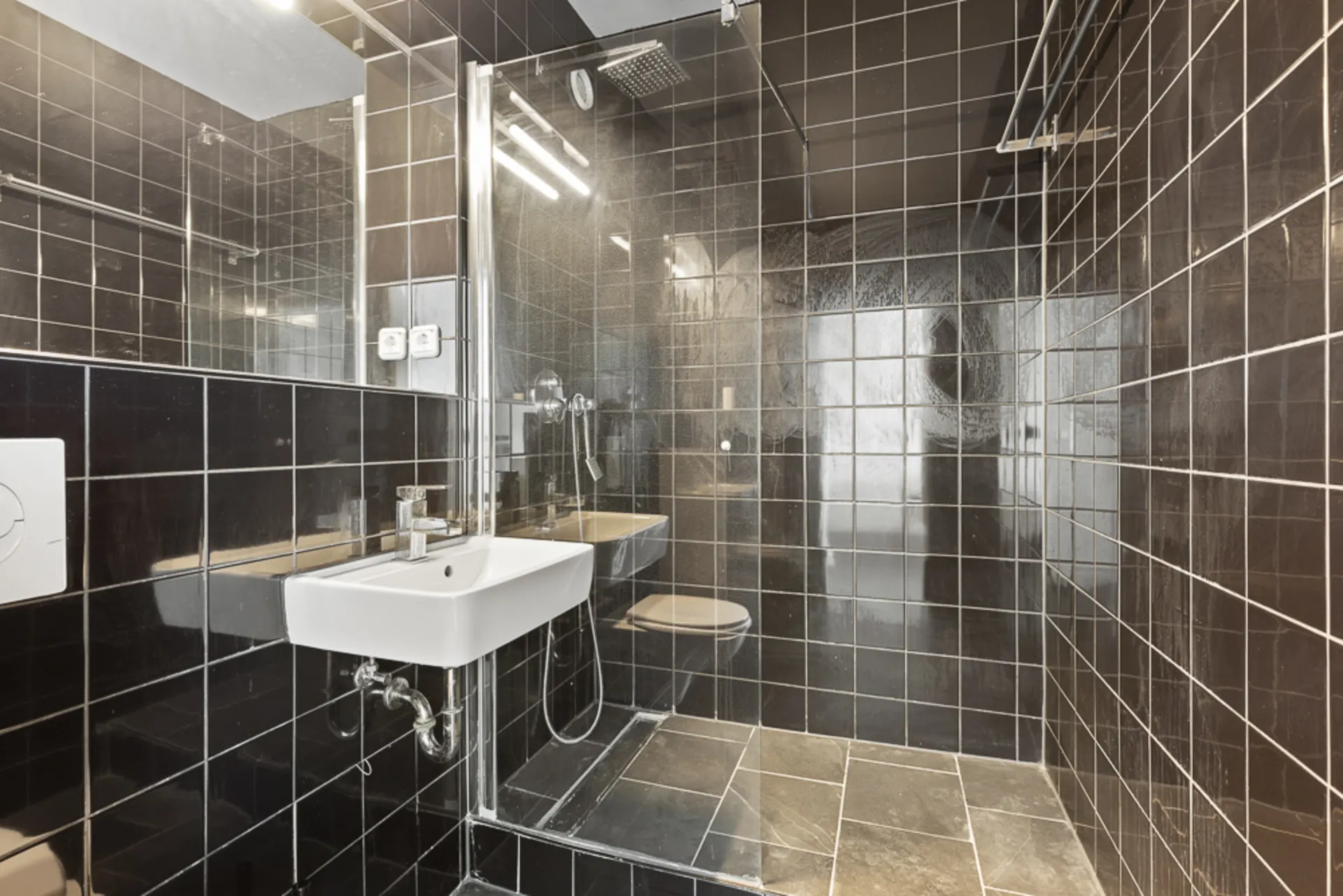 Modernes Badezimmer mit schwarzen Fliesen, Waschbecken, Toilette und offener Dusche.