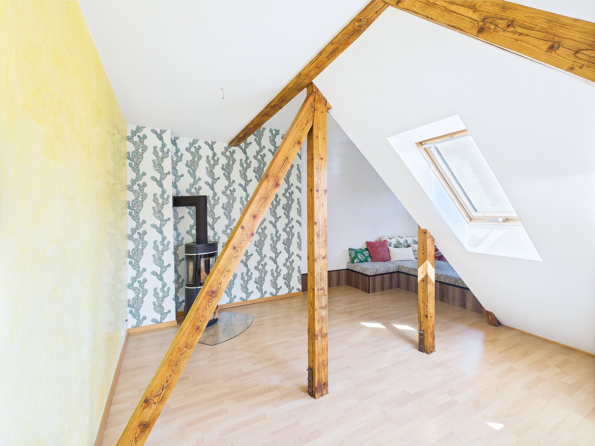 Heller Dachraum mit Holzbalken, Ofen, gelber Wand und Sitzbank unter Dachfenster.