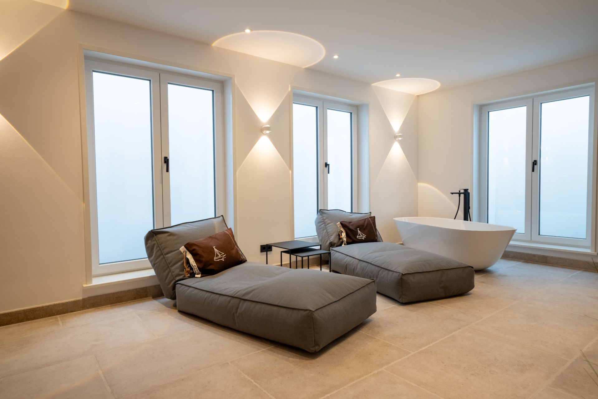 Heller Raum mit zwei grauen Liegesesseln, Badewanne und drei großen Fenstern.