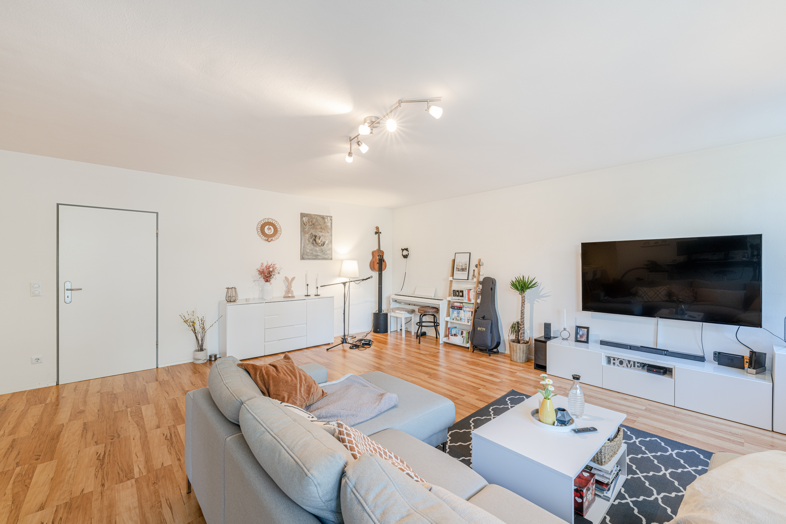 Helles Wohnzimmer mit Sofa, Couchtisch, TV, Regal, Musikinstrumenten und Holzboden.