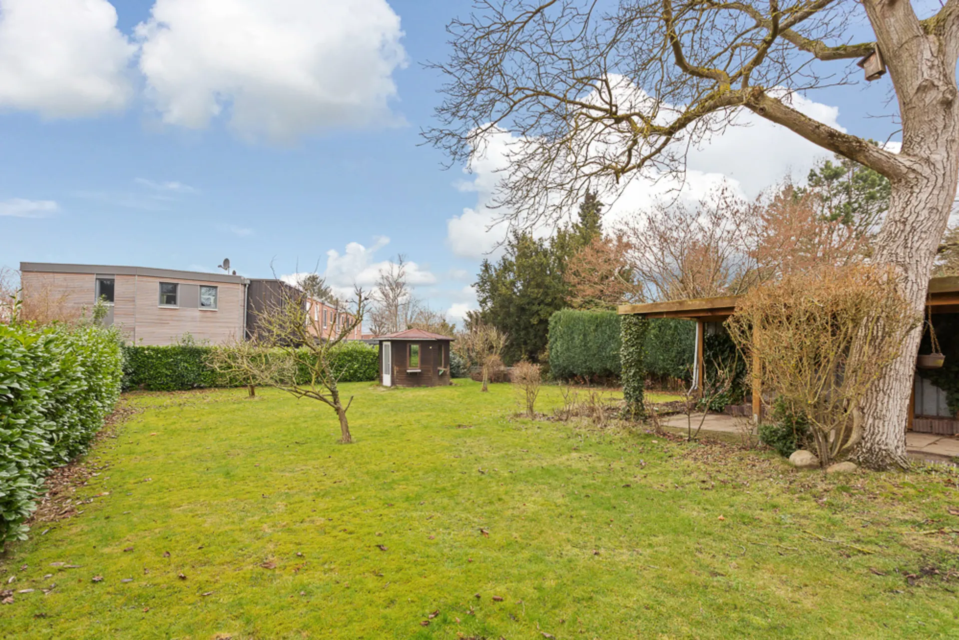 Großer Garten mit Rasen, Bäumen, Gartenhaus und überdachter Terrasse an einem Wohnhaus.