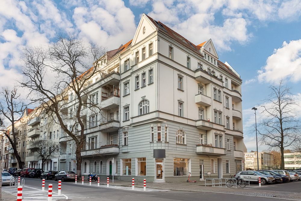 Mehrstöckiges Wohnhaus mit Balkonen an einer Straßenecke, Autos und Bäume am Straßenrand.