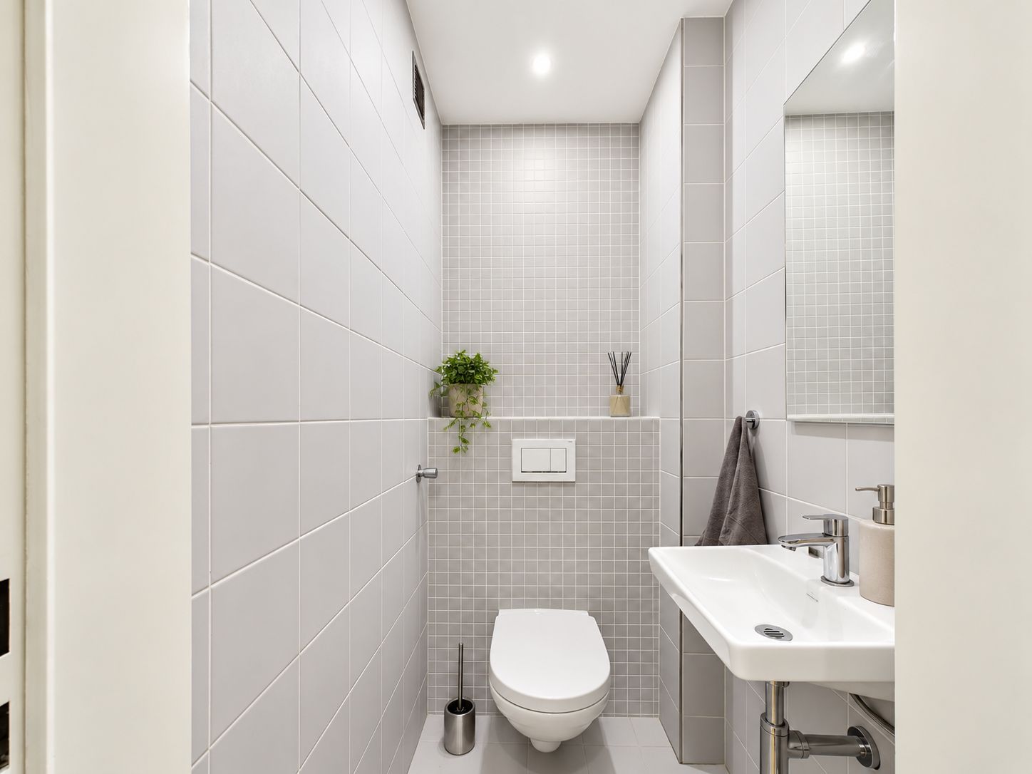Kleines, modernes WC mit Wandtoilette, kleinem Waschbecken und Pflanzen auf einer Ablage.