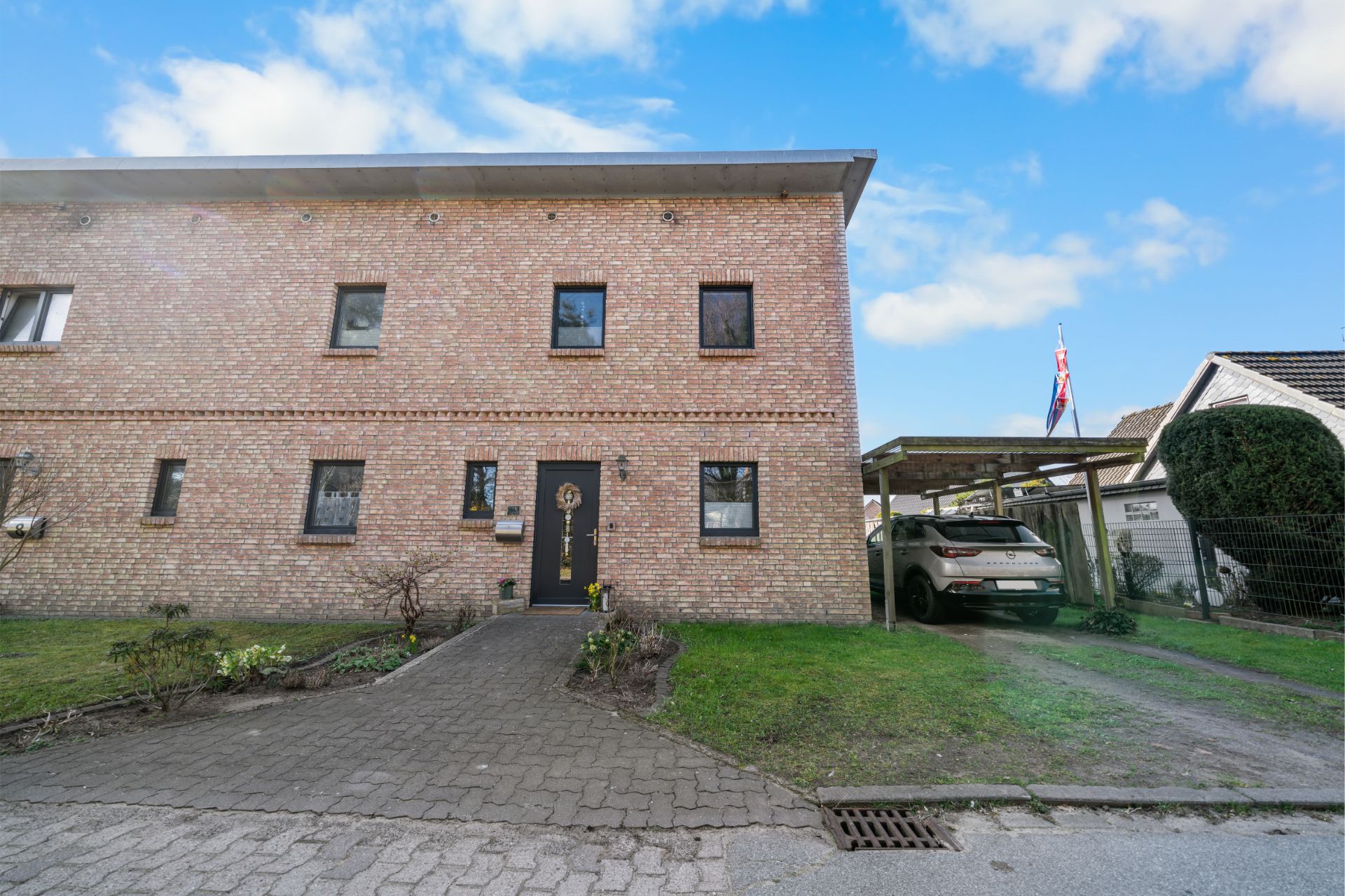 Zweistöckiges Backsteinhaus mit schwarzer Tür, Carport und geparktem Auto rechts daneben.