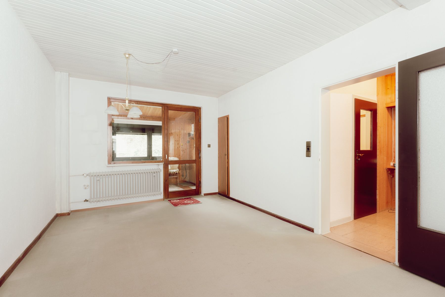 Leerer Raum mit Teppichboden, weißer Decke und Holztür zu einem angrenzenden Zimmer.