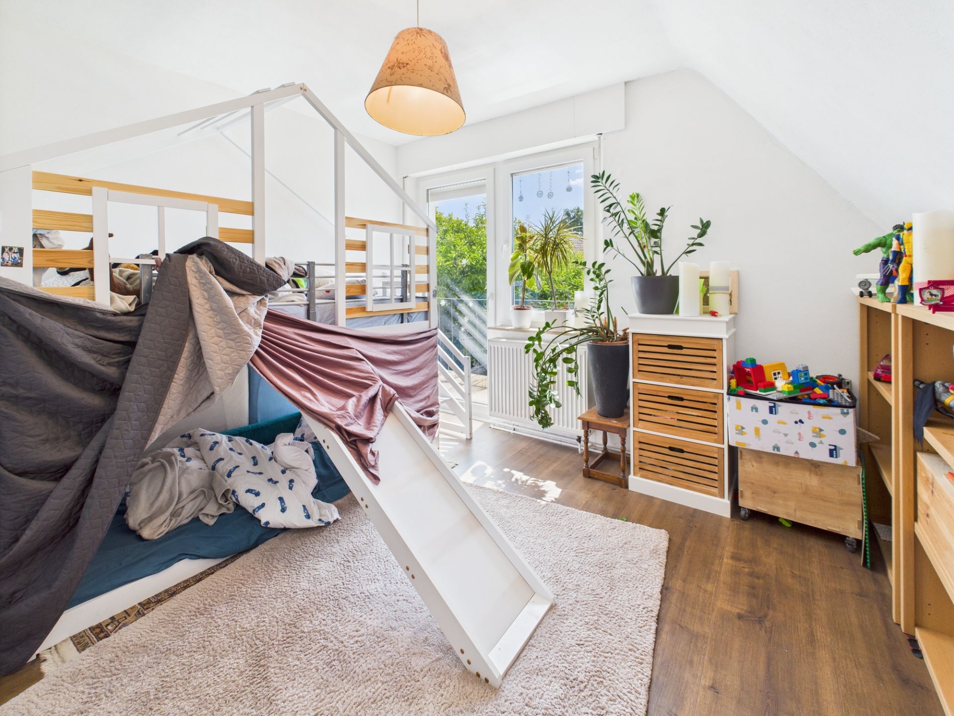 Helles Kinderzimmer mit Hochbett, Rutsche, Spielzeug, Pflanzen und großen Fenstern.